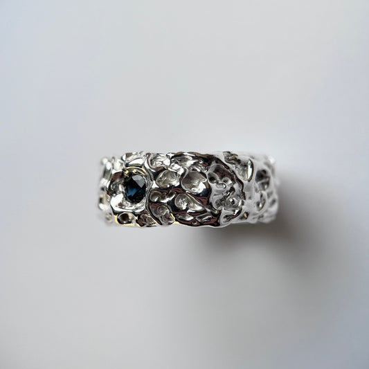 Photo of thick silver ring with one blue/green parti sapphire with an organic texture on a grey background