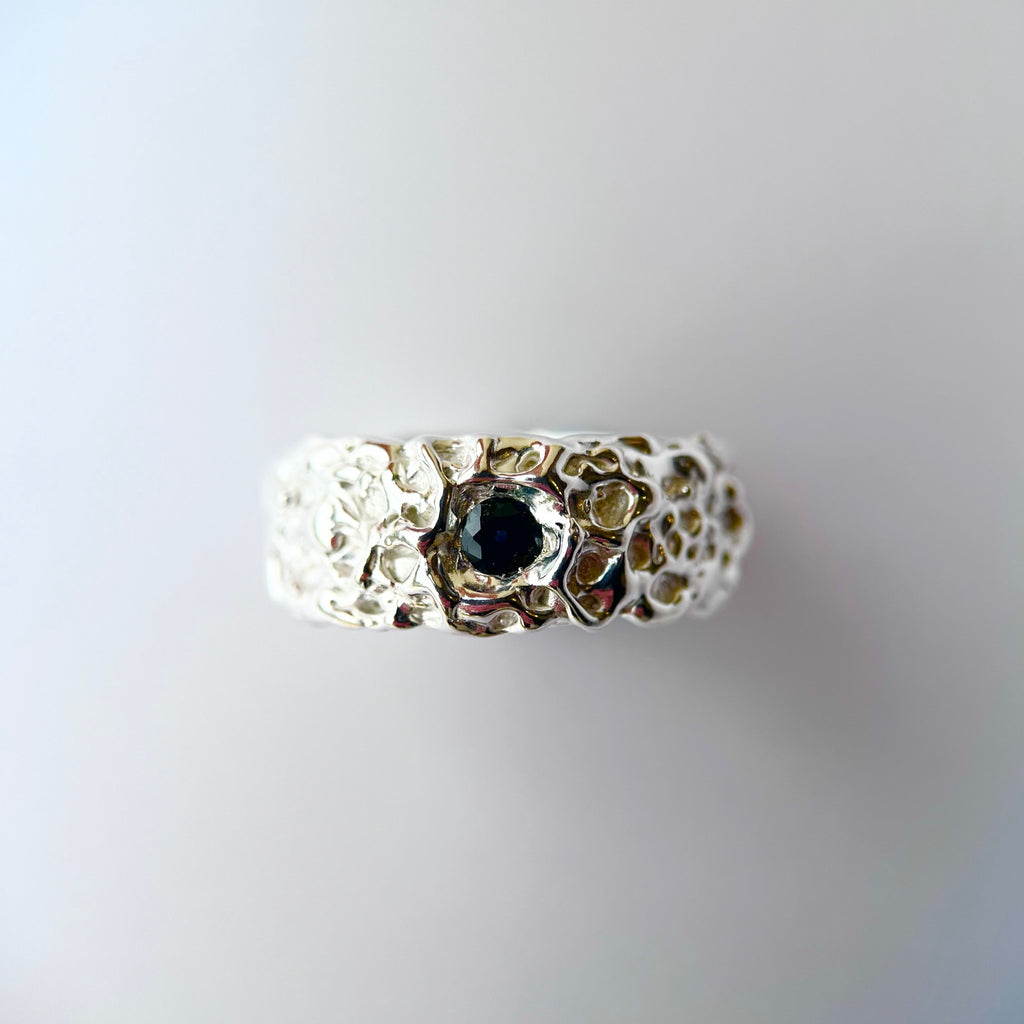 Photo of thick silver band ring with one blue/green parti sapphire with an organic texture on a grey background