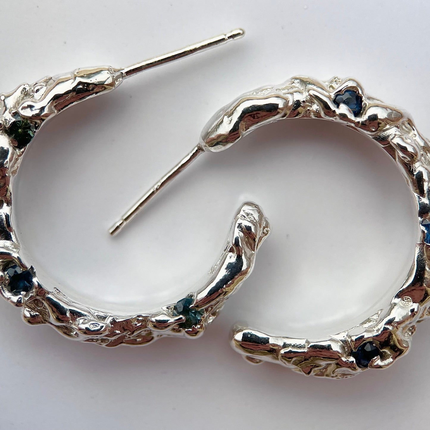 Silver hoop earrings with molten organic texture featuring three blue/green sapphires on each hoop